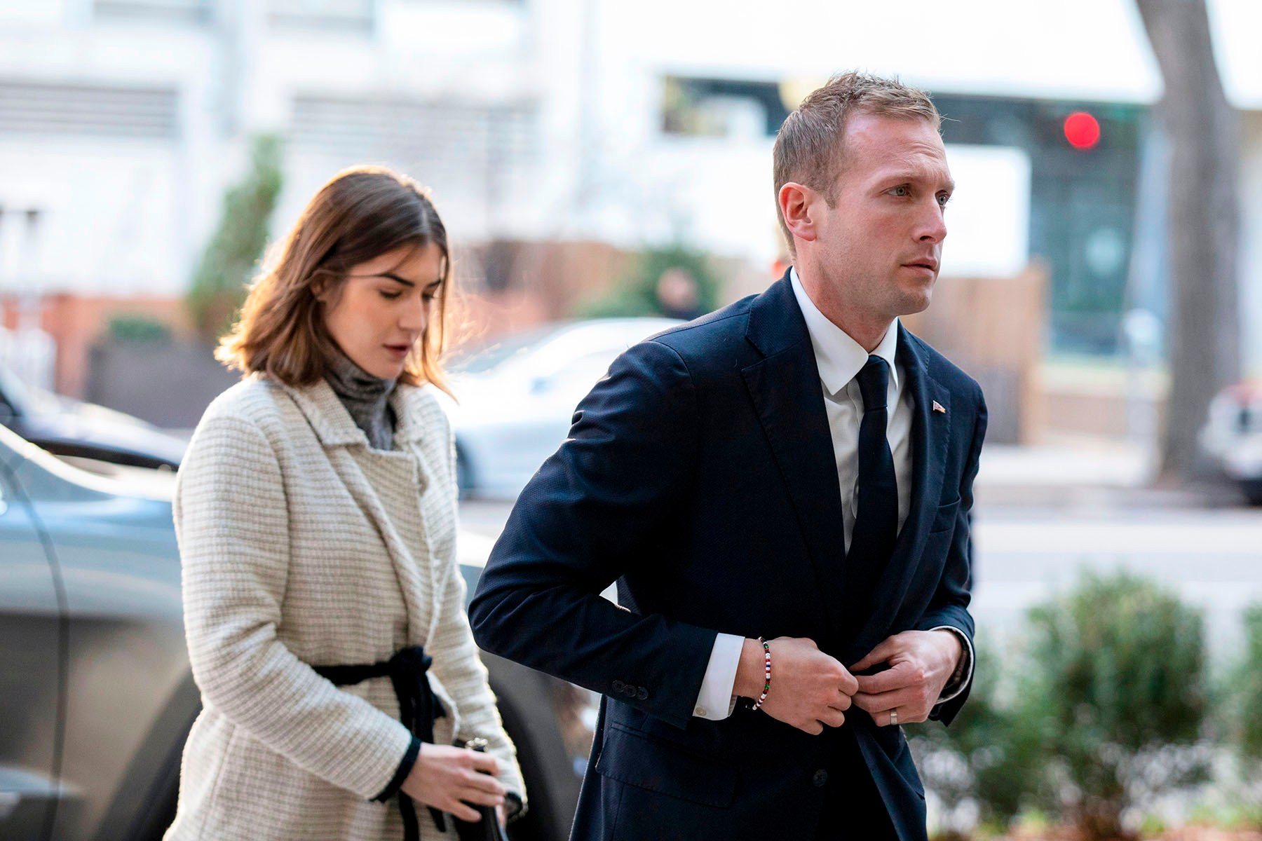 Max Miller, wearing a navy suit and tie, walks alongside Emily Moreno, dressed in a cream-colored coat, outside a building in Washington.