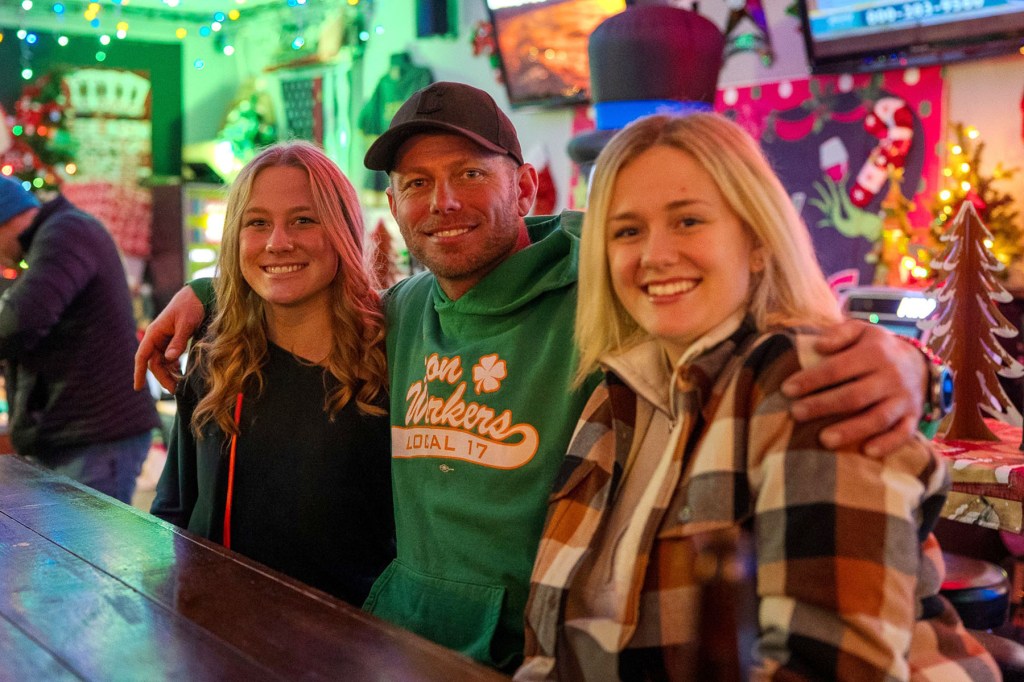 Brian Poindexter, wearing a green Ironworkers Local 17 hoodie and black baseball cap, smiles with his arms around two of his daughters inside a colorful, holiday-decorated bar.