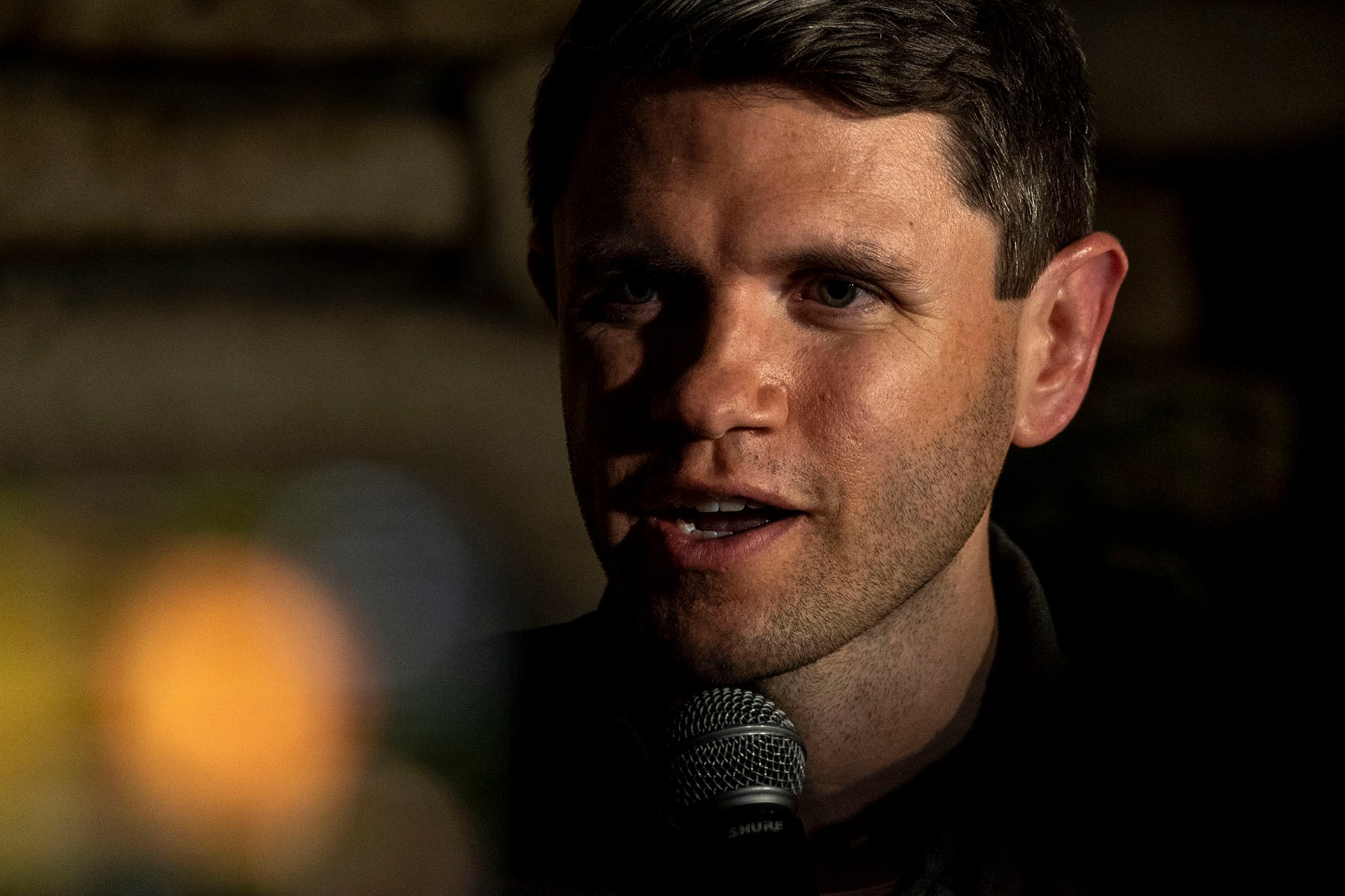 James Talarico speaks into a microphone, his face partially illuminated by warm light against a dark, blurred background.
