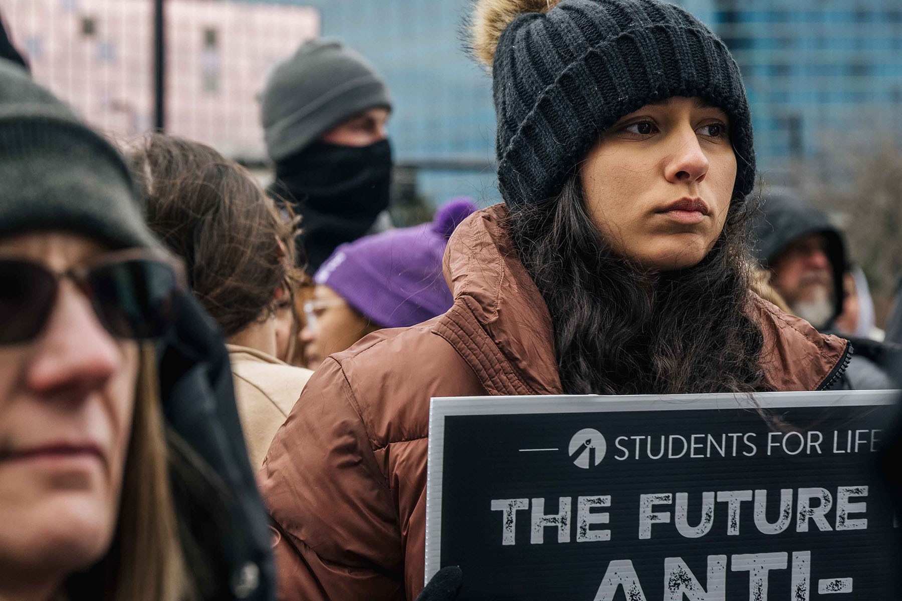 A woman wearing a black knit hat and brown puffer jacket holds a Students for Life anti-abortion sign at a rally.