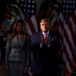 Melania in shadow stands alongside Trump whose face is in a shaft of light as he claps while the background of flags is bathed in shadow.