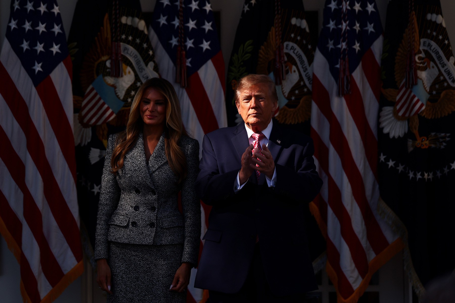 Melania in shadow stands alongside Trump whose face is in a shaft of light as he claps while the background of flags is bathed in shadow.